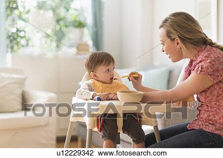 Stock Image - Mother feeding little boy (2-3 years) in high chair. Fotosearch