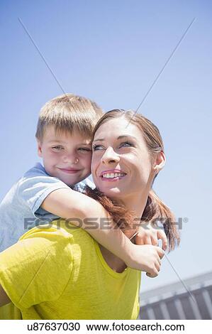 Stock Image - Mother giving her son (6-7) piggyback ride. Fotosearch