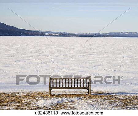 Stock Image - "Park bench at edge of a frozen snowed lake, hills in background". Fotosearch