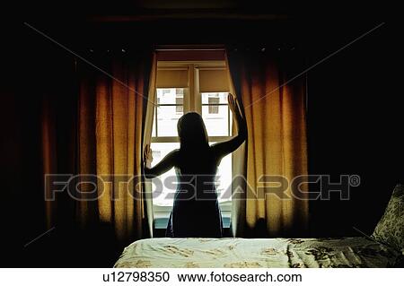 Stock Image - Rear view of female opening curtains at bedroom window. Fotosearch
