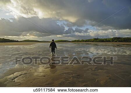 Picture - "Rear view of man standing water in rubber boots, clouds in sky". Fotosearch