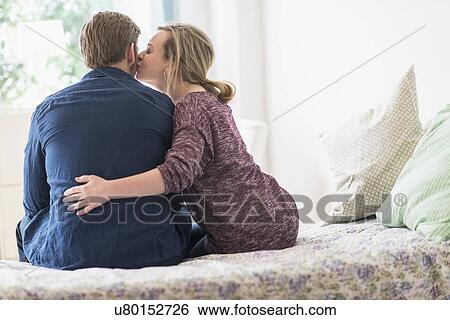 Stock Photograph - Rear view of woman kissing man on cheek on bed. Fotosearch