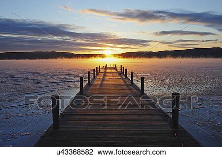Stock Image - "Symmetrical view of jetty on frozen lake, hills in background at sunrise". Fotosearch