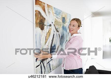 Stock Image - Woman hanging paintings indoors. Fotosearch