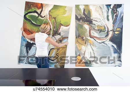 Stock Image - Woman hanging paintings indoors. Fotosearch
