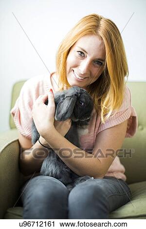 Stock Image - Woman sitting on sofa embracing rabbit. Fotosearch