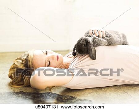 Stock Image - Young woman lying on floor and stroking rabbit. Fotosearch