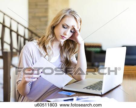 Stock Image - Young woman sitting at table with laptop looking at paper bill. Fotosearch