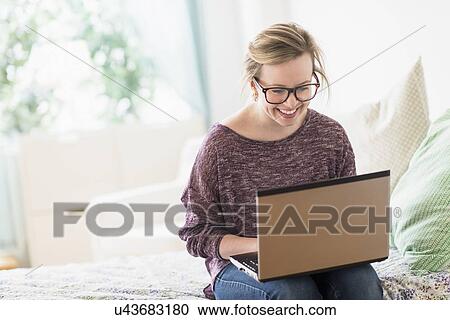 Young woman sitting on bed and using laptop View Large Photo Image Stock Image - Young woman sitting on bed and using laptop. Fotosearch