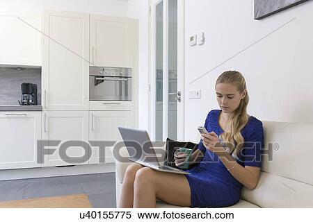 Stock Photography - Young woman using cell phone and laptop at home. Fotosearch
