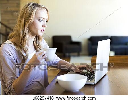 Young woman using laptop and drinking coffee at table View Large Photo Image Stock Photo - Young woman using laptop and drinking coffee at table. Fotosearch