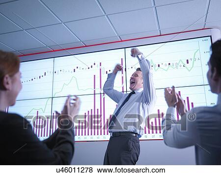 Stock Photo - Businessman celebrating success in front of graphs on screen as colleagues applaud. Fotosearch