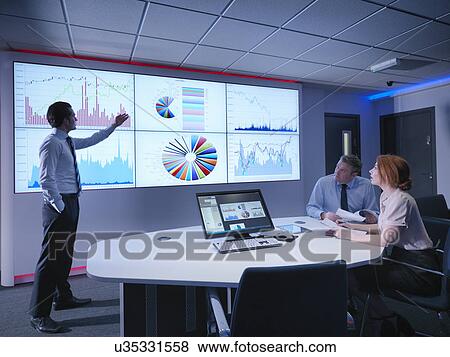 Stock Photo - Businessman making presentation to colleagues in front of graphs on screen. Fotosearch
