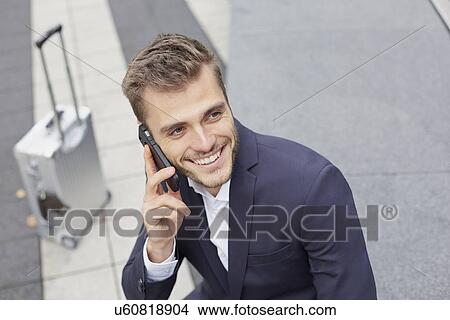 Businessman on sidewalk with wheeled suitcase chatting on smartphone View Large Photo Image Picture - Businessman on sidewalk with wheeled suitcase chatting on smartphone. Fotosearch