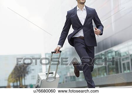 Stock Image - Businessman running with wheeled suitcase outside airport. Fotosearch