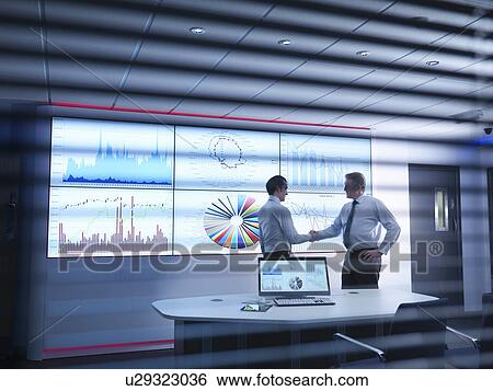 Stock Photograph - Businessmen shaking hands in front of graphs on screen in meeting room. Fotosearch