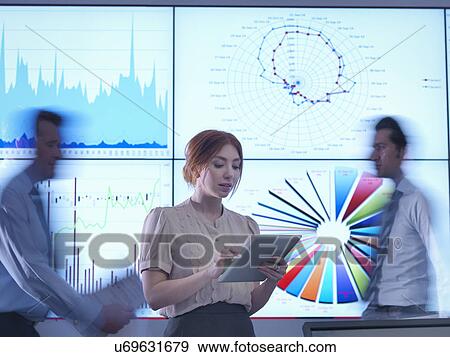 Businesswoman making presentation in front of graphs on screen as colleagues pass behind View Large Photo Image Stock Photo - Businesswoman making presentation in front of graphs on screen as colleagues pass behind. Fotosearch
