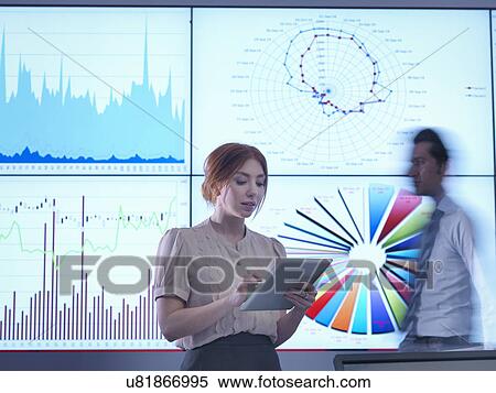 Businesswoman making presentation in front of graphs on screen as colleague passes behind View Large Photo Image Stock Photography - Businesswoman making presentation in front of graphs on screen as colleague passes behind. Fotosearch