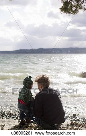 Stock Image - "Father and baby daughter crouching on lakeside, Lake Starnberg, Bavaria, Germany". Fotosearch