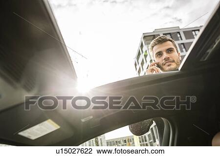Stock Image - Low angle car sunroof view of businessman chatting on smartphone. Fotosearch