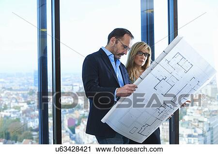 Stock Image - "Male and female architects looking at plans in front of office window with Brussels cityscape, Belgium". Fotosearch