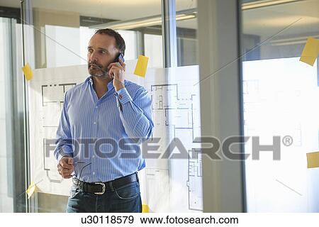 Stock Photo - Male architect using smartphone by plans on glass wall. Fotosearch