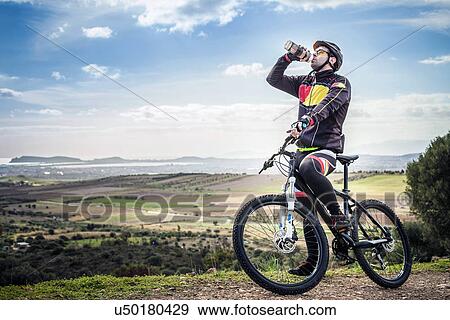 Stock Photo - "Male mountain biker drinking water bottle on coastal path, Cagliari, Sardinia, Italy". Fotosearch