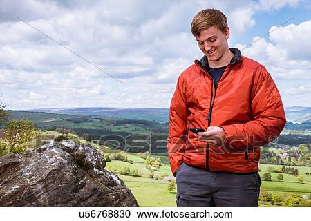 Stock Image - "Male teenage hiker reading smartphone texts on top of Guise Cliff, Pateley Bridge, Nidderdale, Yorkshire Dales". Fotosearch