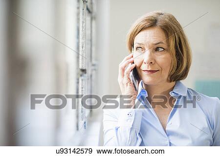 Stock Photo - Mature businesswoman looking out of office window and chatting on smartphone. Fotosearch