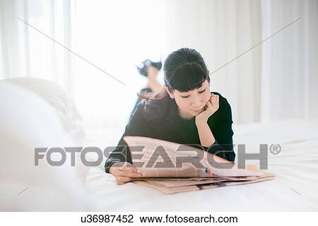 Stock Image - Mature businesswoman lying on bed reading newspaper in hotel room. Fotosearch