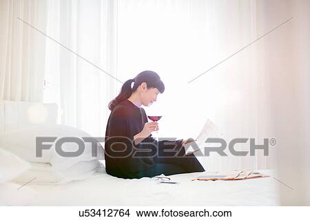 Picture - Mature businesswoman reading newspaper whilst drinking red wine in hotel room. Fotosearch