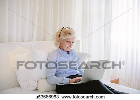 Mature businesswoman sitting on bed typing on laptop in hotel room View Large Photo Image Stock Image - Mature businesswoman sitting on bed typing on laptop in hotel room. Fotosearch