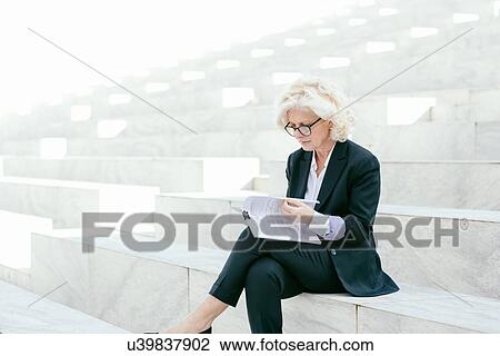 Stock Image - "Mature businesswoman sitting on steps, looking through documents". Fotosearch