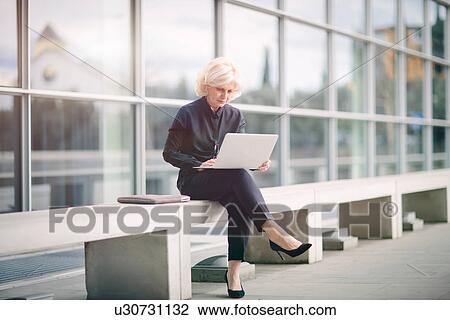 "Mature businesswoman, sitting on bench, outside building, using laptop" View Large Photo Image Stock Image - "Mature businesswoman, sitting on bench, outside building, using laptop". Fotosearch