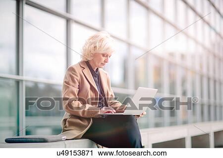 Stock Image - "Mature businesswoman, sitting on bench, outside building, using laptop". Fotosearch