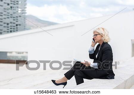 Stock Image - "Mature businesswoman sitting on wall, using mobile phone, holding digital tablet". Fotosearch