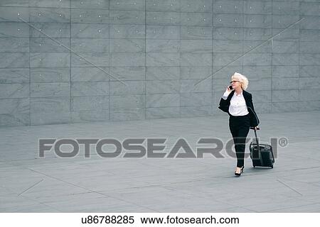 "Mature businesswoman walking with suitcase, using mobile phone" View Large Photo Image Stock Photography - "Mature businesswoman walking with suitcase, using mobile phone". Fotosearch
