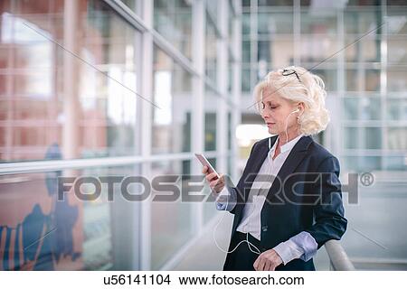 "Mature businesswoman wearing earphones, looking at smartphone, standing in corridor" View Large Photo Image Picture - "Mature businesswoman wearing earphones, looking at smartphone, standing in corridor". Fotosearch