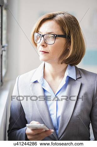 Mature businesswoman with smartphone gazing out of office window View Large Photo Image Stock Photograph - Mature businesswoman with smartphone gazing out of office window. Fotosearch