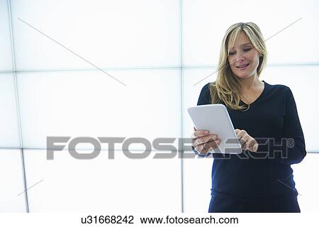 Stock Image - Mature woman using digital tablet against backlit wall panels. Fotosearch