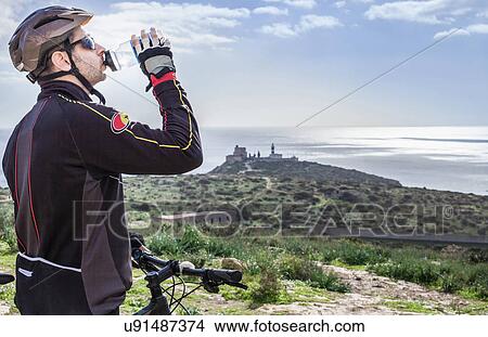 "Mid adult male mountain biker drinking water on coastal path, Cagliari, Sardinia, Italy" View Large Photo Image Picture - "Mid adult male mountain biker drinking water on coastal path, Cagliari, Sardinia, Italy". Fotosearch