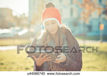 Stock Image - Mid adult woman in red pom pom hat using digital tablet in park. Fotosearch