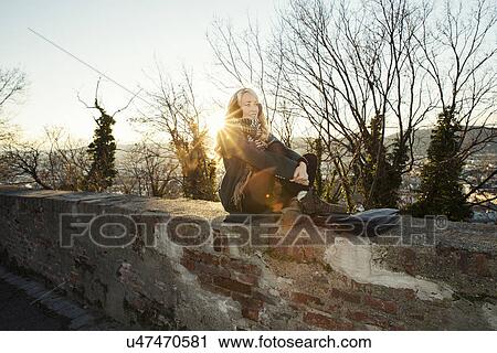 Stock Image - Mid adult woman sitting on wall in sunlight. Fotosearch