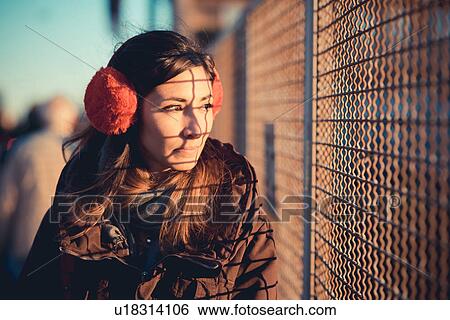 Stock Photograph - Mid adult woman wearing red ear muffs gazing through sunlit wire fence. Fotosearch