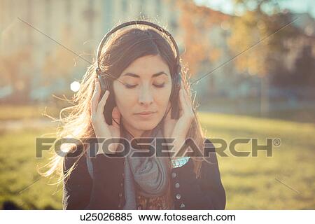 Stock Photography - Mid adult woman with eyes closed listening to headphones in park. Fotosearch