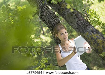 Stock Image - Portrait of young woman leaning on tree branch using digital tablet touchscreen. Fotosearch