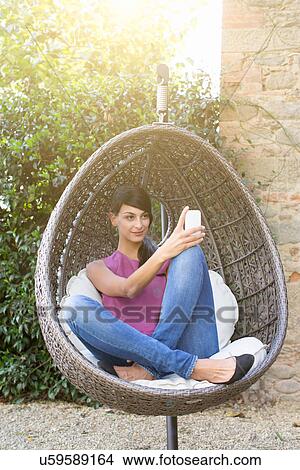 Portrait of young woman sitting in swing chair using smartphone in garden View Large Photo Image Picture - Portrait of young woman sitting in swing chair using smartphone in garden. Fotosearch
