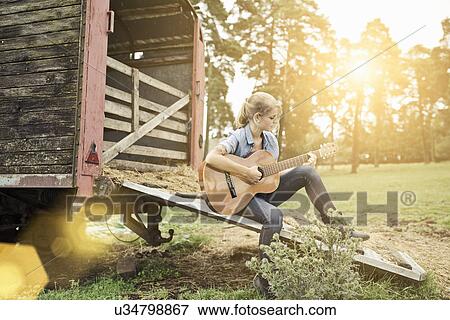 Stock Photo - "Portrait of young woman, sitting on wooden cart, playing guitar". Fotosearch