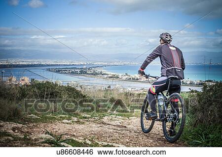 Stock Photograph - "Rear view of male mountain biker looking out from coastal path, Cagliari, Sardinia, Italy". Fotosearch