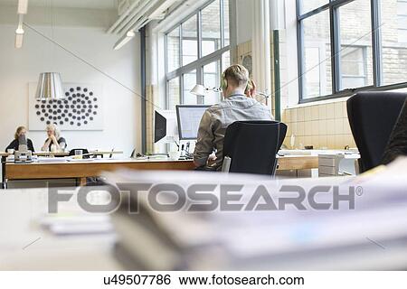 Stock Photograph - Rear view of man working at desk whilst listening to headphones. Fotosearch